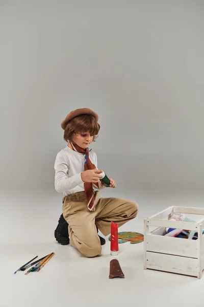 Niño se arrodilla en el suelo, rodeado de pinturas en tubos y una caja de herramientas de madera, joven pintor en boina - foto de stock