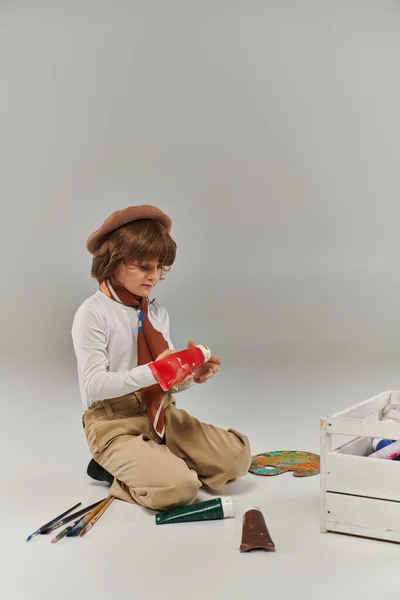 Niño se arrodilla en el suelo, rodeado de pinturas en tubos y una caja de herramientas de madera, joven artista en boina - foto de stock