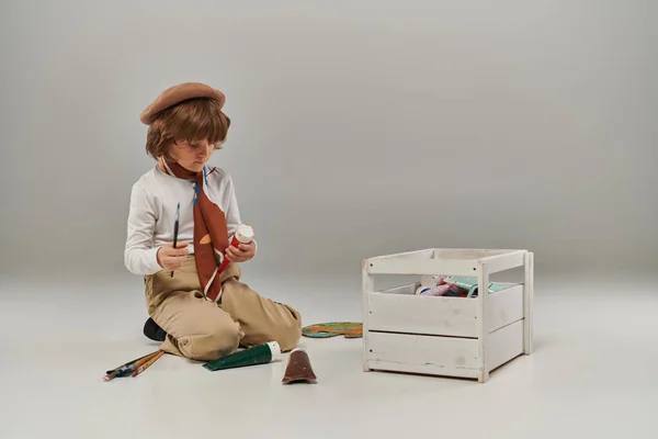 Niño se arrodilla en el suelo rodeado de pinturas en tubos y una caja de herramientas de madera, joven artista en boina - foto de stock