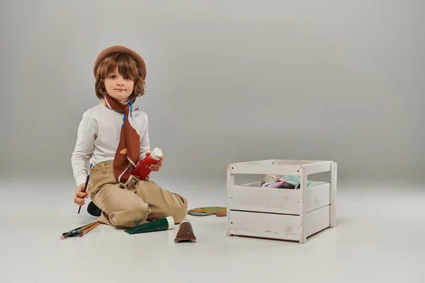Niño se arrodilla en el suelo rodeado de pinturas acrílicas en tubos y caja de madera, joven artista en boina - foto de stock