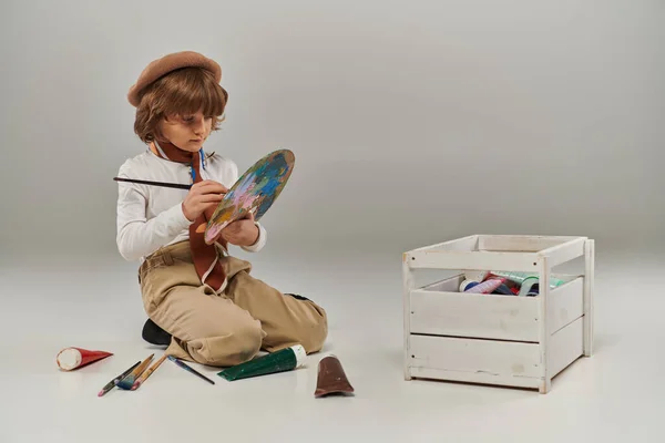 Young artist mixing paint on palette, boy in beret surrounded by paint brushes and colorful tubes — Stock Photo