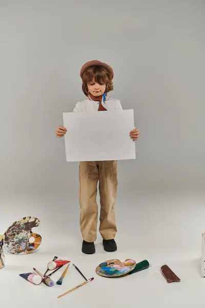 Young artist holding blank paper near paint brushes and palette on floor, boy in beret and scarf — Stock Photo