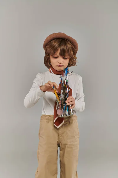 Joven artista sosteniendo frasco de vidrio con pinceles sobre fondo gris, niño en boina y bufanda - foto de stock
