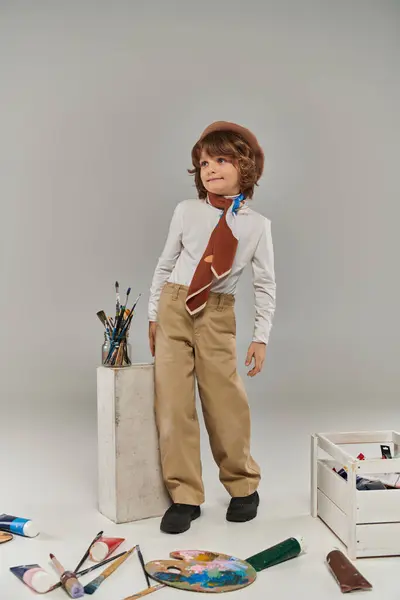 Artiste élégant debout près de bloc blanc avec pinceaux, garçon en béret et écharpe en studio — Photo de stock