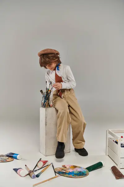Artiste élégant assis près bloc blanc avec pinceaux, garçon en béret et écharpe en studio — Photo de stock