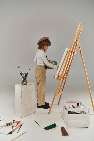 Молодий художник з палітрою і розписом пензлем біля мольберта, стильний хлопчик в береті і шарфі — стокове фото