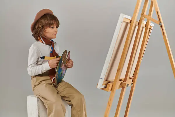 Felice ragazzo esplora il suo potenziale creativo, bambino in berretto con tavolozza guardando cavalletto con tela — Foto stock