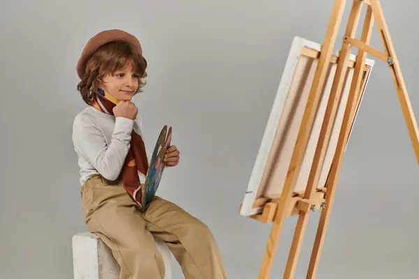 Веселый молодой художник раскрывает свой творческий потенциал, мальчик в берете смотрит на мольберт с холстом — стоковое фото