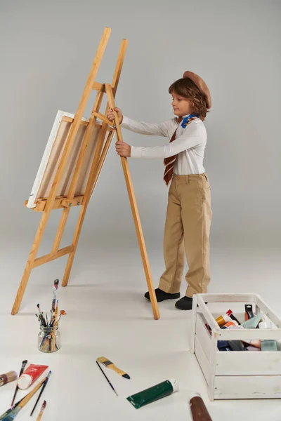Young artist adjusting his easel, surrounded by his tools and colorful paint, boy in beret and scarf — Stock Photo