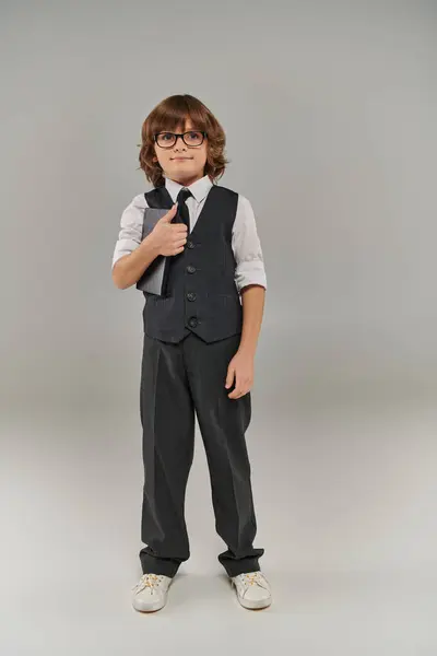 Ein kleiner Junge in modischer Weste und Krawatte hält ein Buch in der Hand und steht auf grauem Hintergrund — Stockfoto