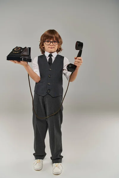 Élégant garçon dans des lunettes et gilet élégant tenant téléphone rétro sur fond gris, nostalgie — Photo de stock