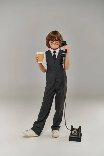Garçon élégant dans des lunettes et élégant costume formel tenant téléphone rétro et boisson en tasse de papier sur gris — Photo de stock