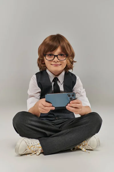 Улыбающийся мальчик в очках и элегантный наряд, играющий в мобильную игру на сером, держа смартфон — стоковое фото