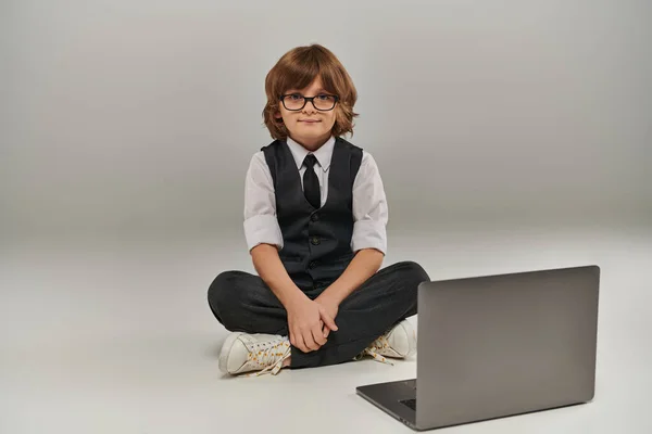 Boy in elegant attire with vest and trousers sitting near laptop on grey, future businessman — Stock Photo