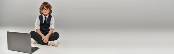 Boy in elegant attire with vest and trousers sitting near laptop on grey, future businessman, banner — Stock Photo