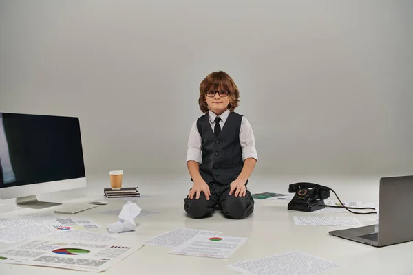 Menino de óculos cercado por papéis e material de escritório em pano de fundo cinza, futuro empresário — Fotografia de Stock