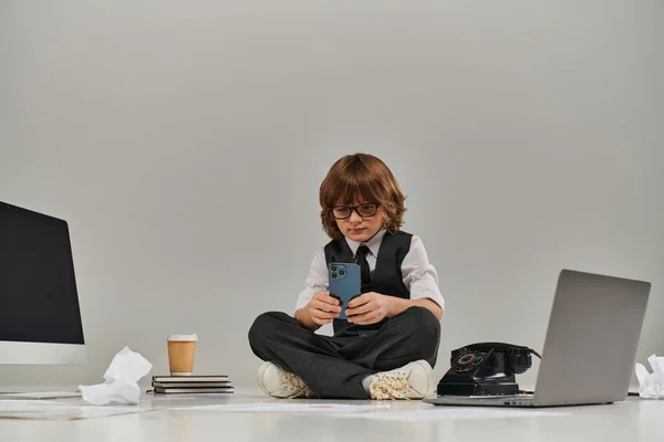 Niño sentado en el suelo, absorto en la tecnología como él utilizando tanto el teléfono como el ordenador portátil, futuro hombre de negocios - foto de stock