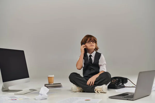 Niño en gafas y atuendo formal hablando en smartphone y sentado rodeado de equipo de oficina - foto de stock