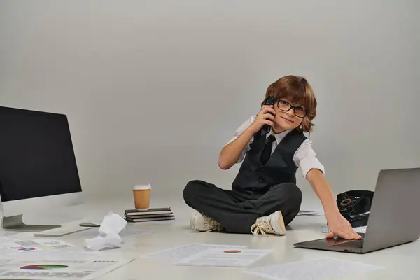 Niño en gafas y ropa formal hablando en smartphone y sentado rodeado de equipo de oficina - foto de stock