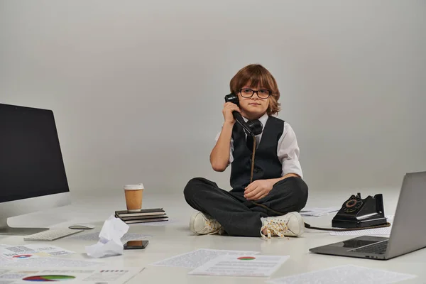 Niño en gafas y ropa formal hablando por teléfono retro y sentado rodeado de equipo de oficina - foto de stock