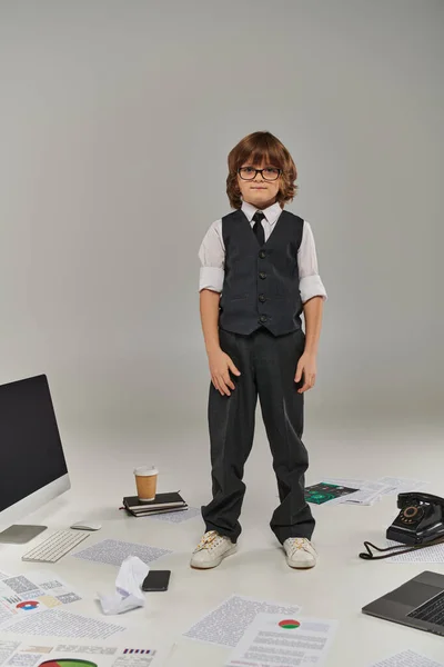 Kid in glasses and formal wear surrounded by office equipment and devices standing on grey — Stock Photo