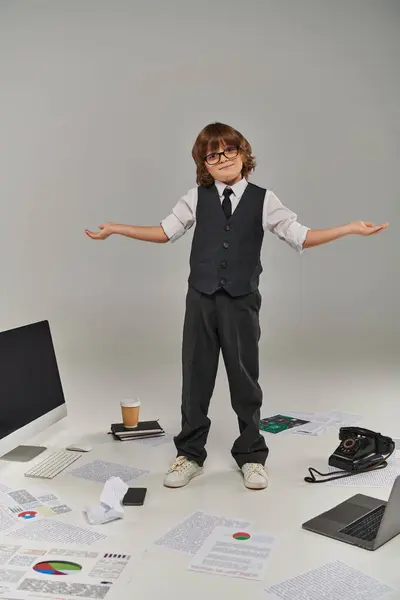 Confused kid in glasses and formal wear surrounded by office equipment and devices standing on grey — Stock Photo