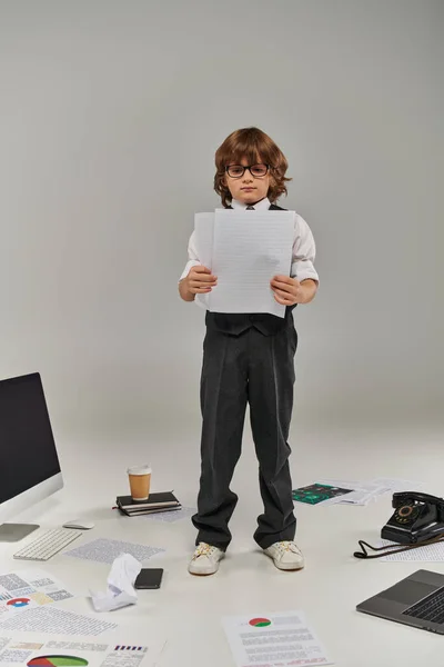 Мила дитина в окулярах і формальний одяг в оточенні офісного обладнання і пристроїв, що стоять з паперами — стокове фото