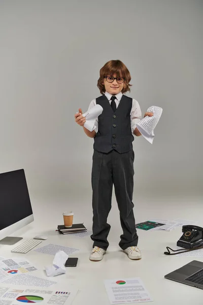 Cute boy in glasses and formal wear surrounded by office equipment and devices standing with papers — Stock Photo