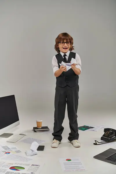 Excited boy in glasses and formal wear surrounded by office equipment and devices holding papers — Stock Photo