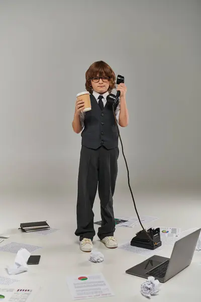 Lindo chico en gafas y formal desgaste sosteniendo taza de papel y teléfono retro durante la llamada, multitarea - foto de stock