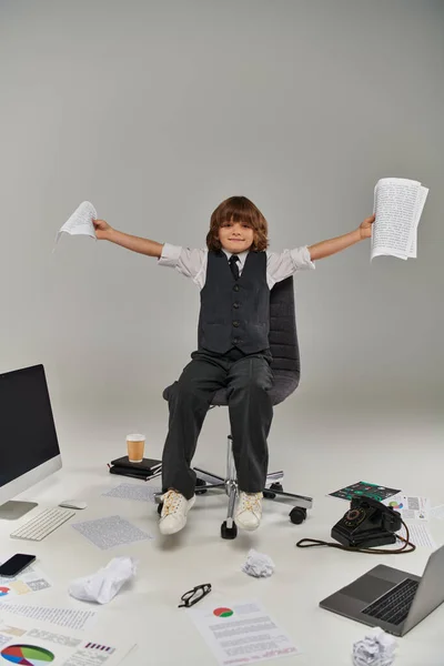 Menino feliz com papéis nas mãos sentado na cadeira cercado por material de escritório, futuro profissional — Fotografia de Stock