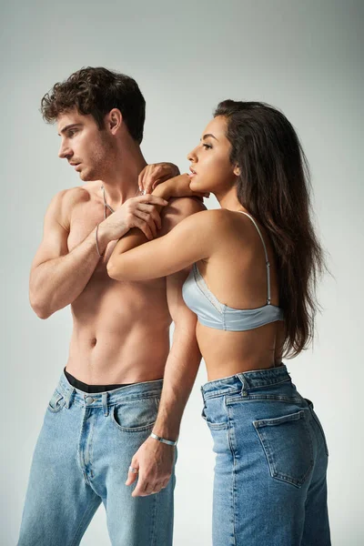 Beautiful young woman in satin bra and jeans embracing shirtless man on grey background, affection — Stock Photo