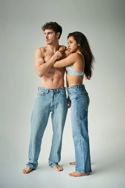 Brunette young woman in satin bra and jeans embracing shirtless man on grey background, affection — Stock Photo