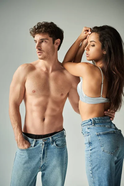 Attraente giovane donna in reggiseno di raso e jeans abbracciando uomo senza camicia su sfondo grigio, affetto — Foto stock