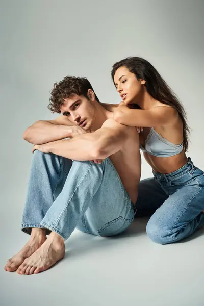Сексуальная молодая женщина в атласном лифчике и джинсах позирует с мужчиной без рубашки на сером фоне, близость — стоковое фото