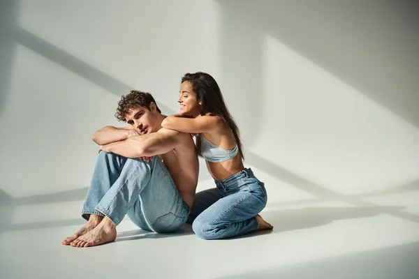 Jovem feliz em sutiã de cetim e jeans posando com homem sem camisa no fundo cinza, proximidade — Fotografia de Stock