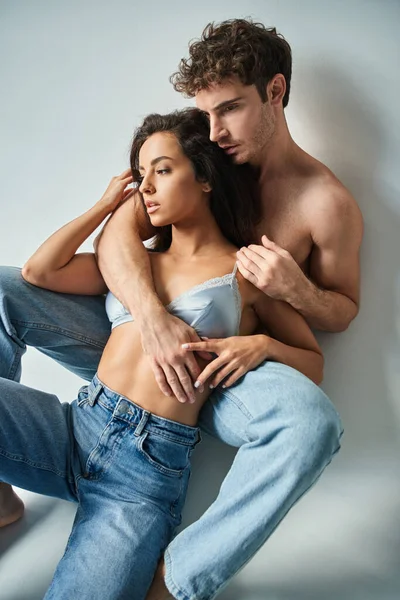 Passionate and shirtless man in jeans sitting and hugging brunette girlfriend in bra on grey — Stock Photo