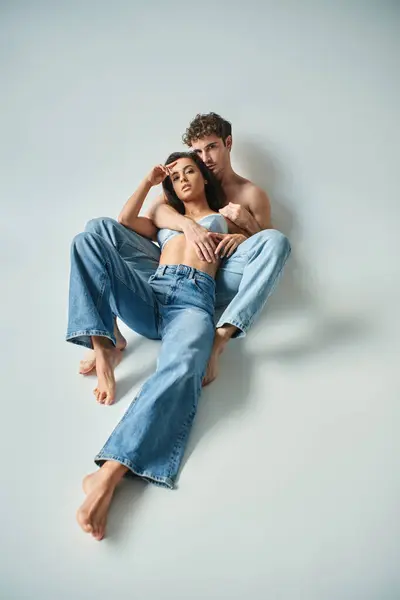 Passionné et pieds nus homme en jeans assis et câlin petite amie brune en soutien-gorge sur gris — Photo de stock