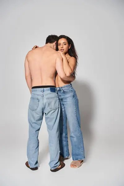 Sexy y sin camisa hombre abrazando a mujer joven en vaqueros vaqueros azul sobre fondo gris, juntos - foto de stock