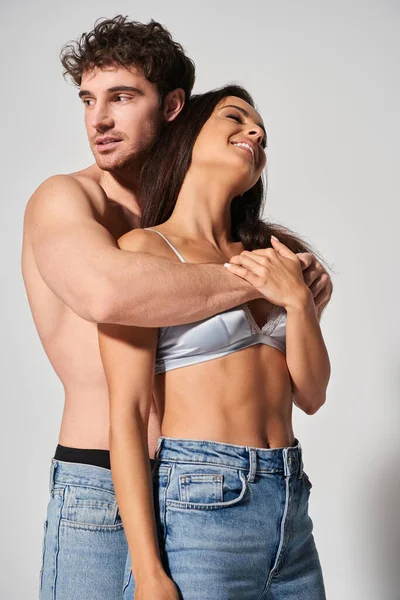 Sexy e senza maglietta uomo abbracciare seducente giovane e allegra donna su sfondo grigio, insieme — Foto stock