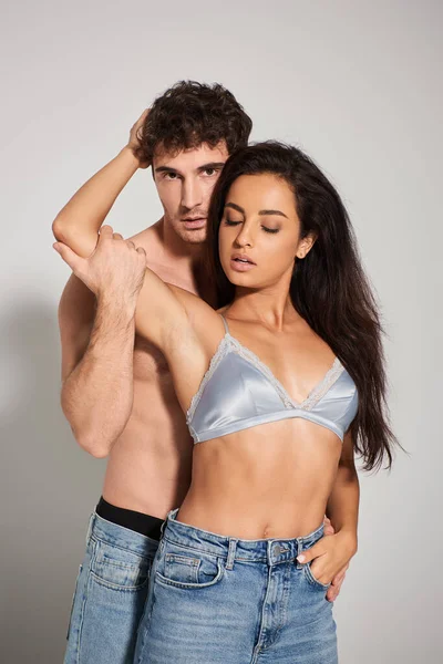 Seductive young woman with brunette hair hugging shirtless man on grey background, vertical — Stock Photo