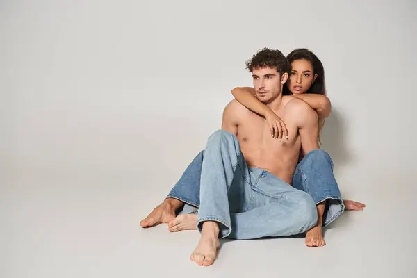 Barfuß und sexy Frau in blauen Jeans umarmt ihren hemdlosen Freund auf grauem Hintergrund, Liebe — Stockfoto