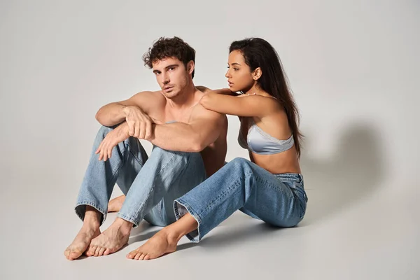 Brunette young woman in blue jeans embracing shirtless man on grey background, full length — Stock Photo