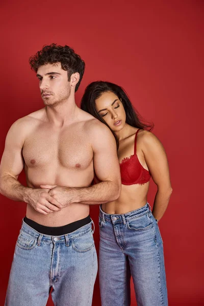 Sensual joven mujer en sujetador y vaqueros apoyados en el hombre sin camisa sobre fondo rojo, afecto - foto de stock