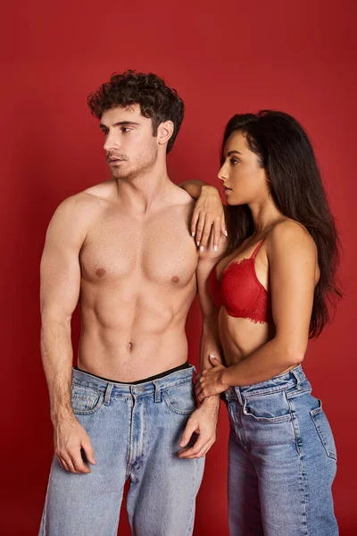 Sensual young woman with brunette hair leaning on body of muscular man on red background, affection — Stock Photo