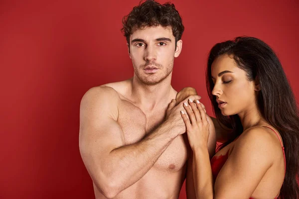 Homme musclé et torse nu regardant la caméra près de magnifique femme brune en soutien-gorge sur fond rouge — Photo de stock