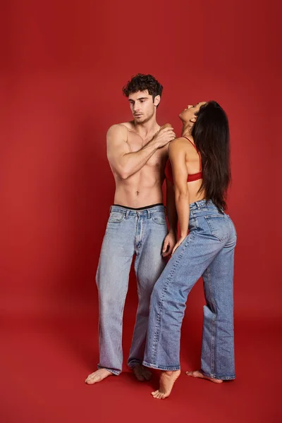 Homme musclé et torse nu en jeans debout près de magnifique femme brune en soutien-gorge sur fond rouge — Photo de stock