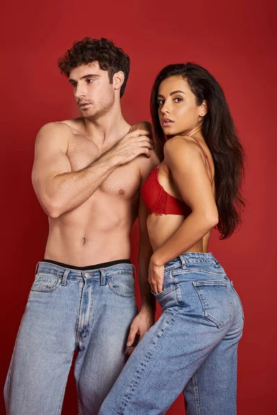 Homme musclé et torse nu en jeans debout avec magnifique femme brune en soutien-gorge sur fond rouge — Photo de stock