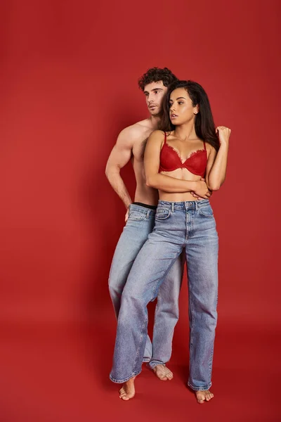 Bel homme torse nu en jeans posant avec belle femme en lingerie sexy sur fond rouge — Photo de stock