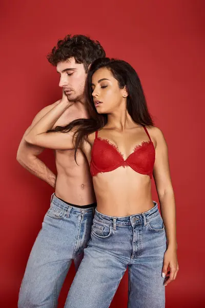 Bela jovem mulher no sexy lingerie abraçando seu sexy muscular namorado no fundo vermelho — Fotografia de Stock